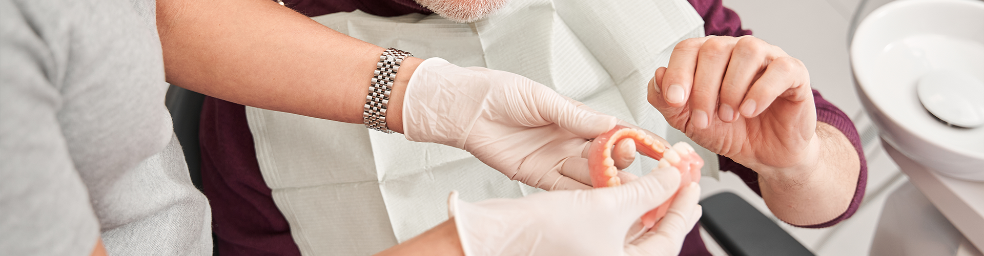 gloved hands handing dentures to man wearing bib in dental chair.