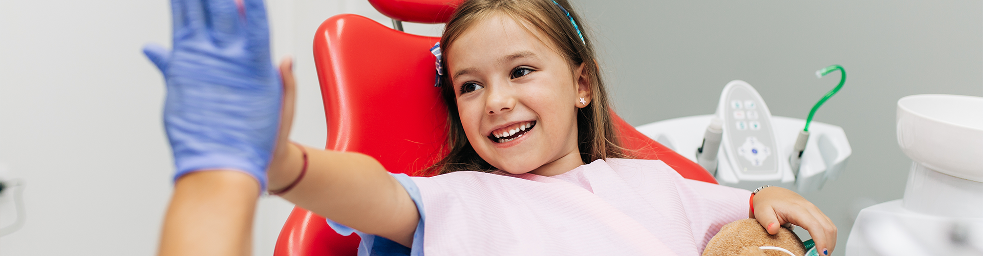 gloved hand high-fiving smiling child in red dental chair.